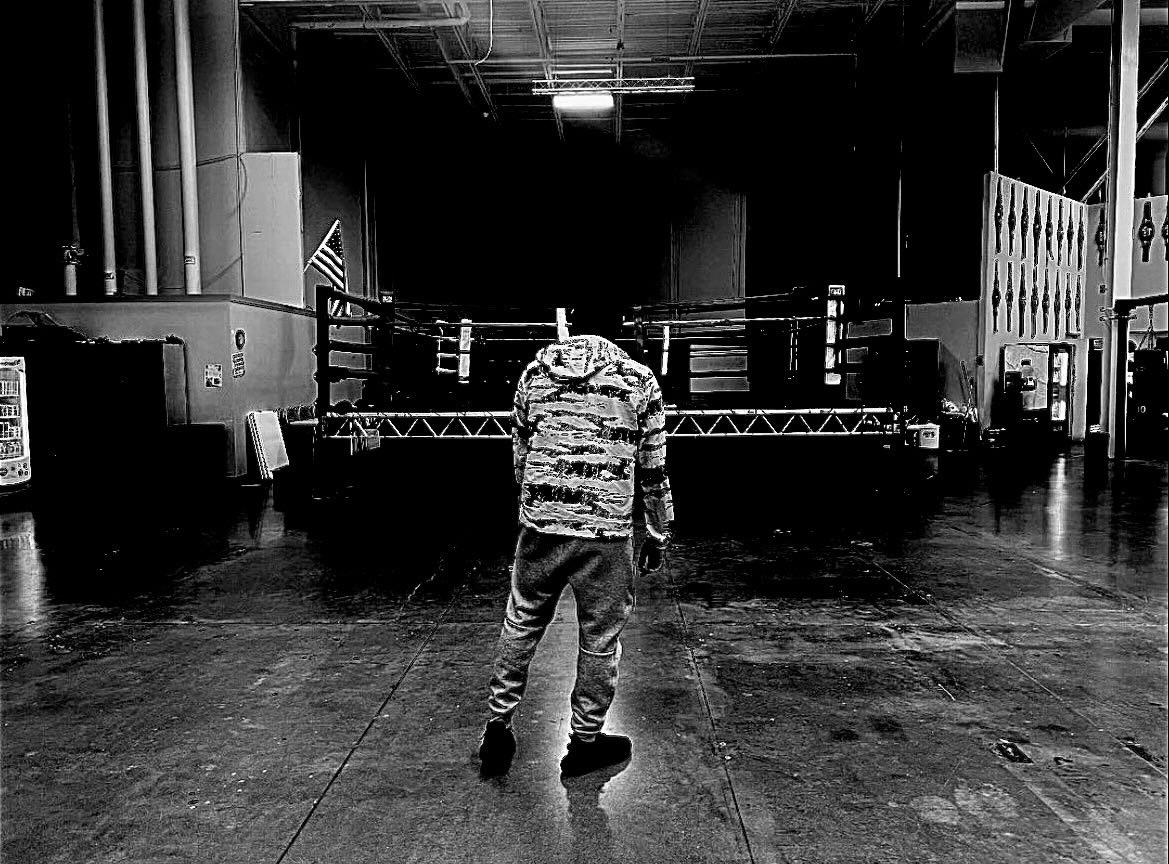 Black-and-white photo of a hooded figure standing alone in front of a boxing ring